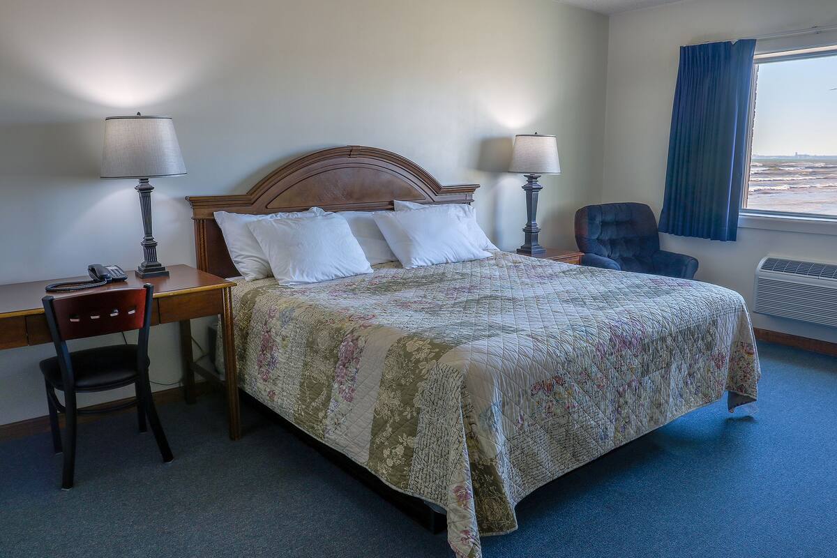 A hotel room with a bed, two bedside lamps, a desk with a chair, and a view of the ocean through a window.