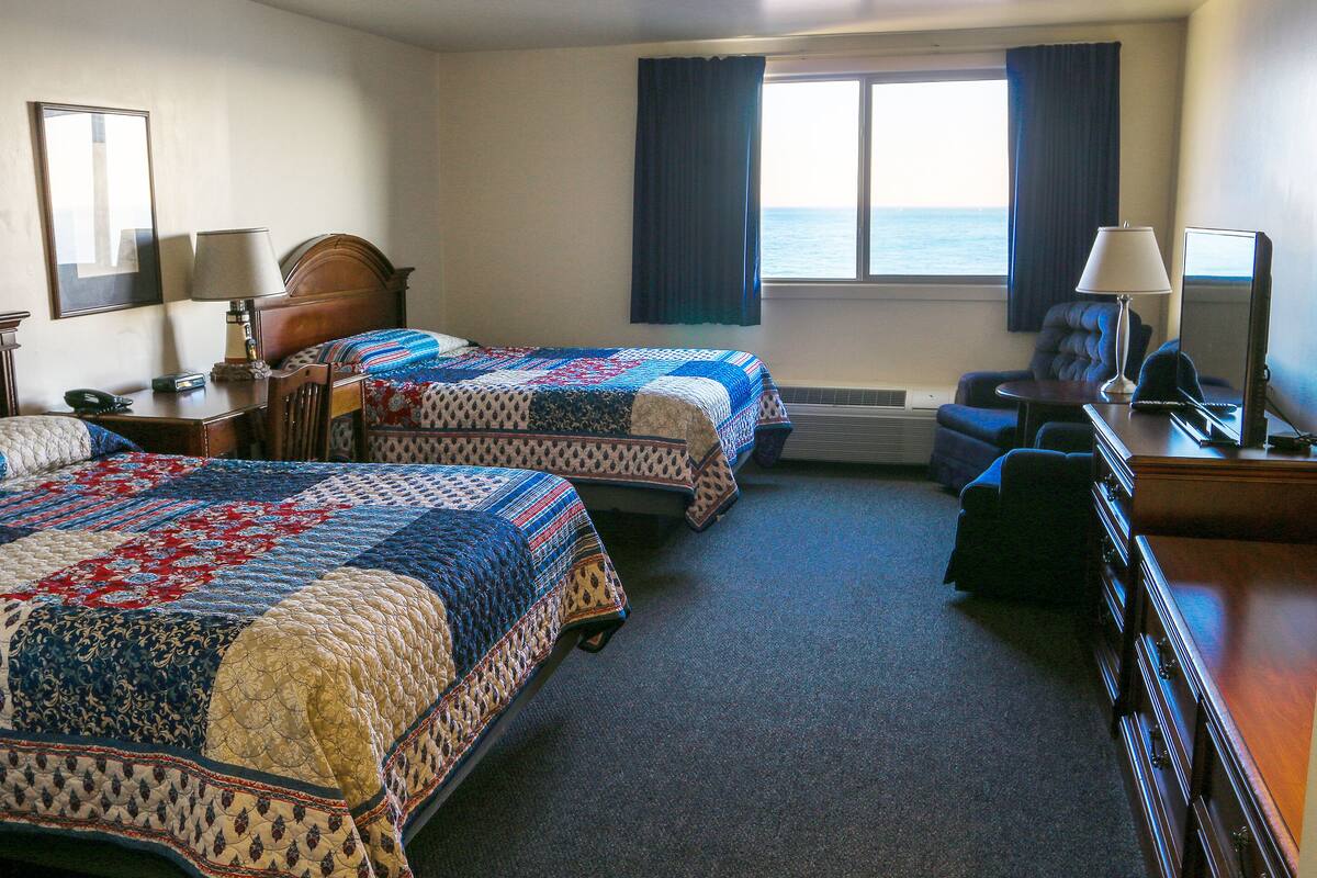 A hotel room with two beds, a desk, a chair, a TV, and a view of the sea.