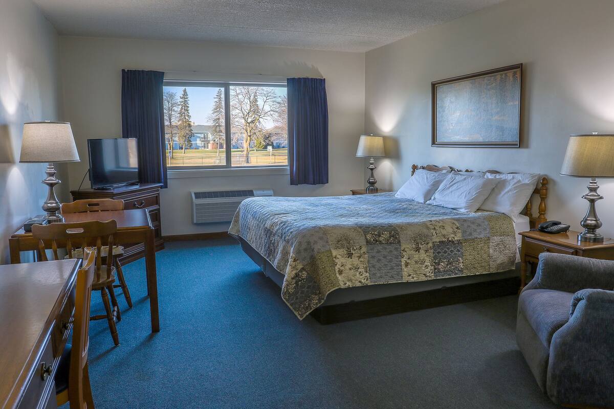 A hotel room with a bed, a desk with a chair, a television, a window with curtains, and a painting on the wall.