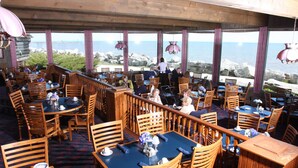 A restaurant with wooden tables and chairs, large windows with a view of the sea, and hanging pendant lights.