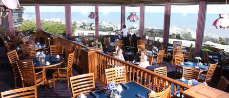 A restaurant with wooden tables and chairs, large windows with a view of the sea, and hanging pendant lights.