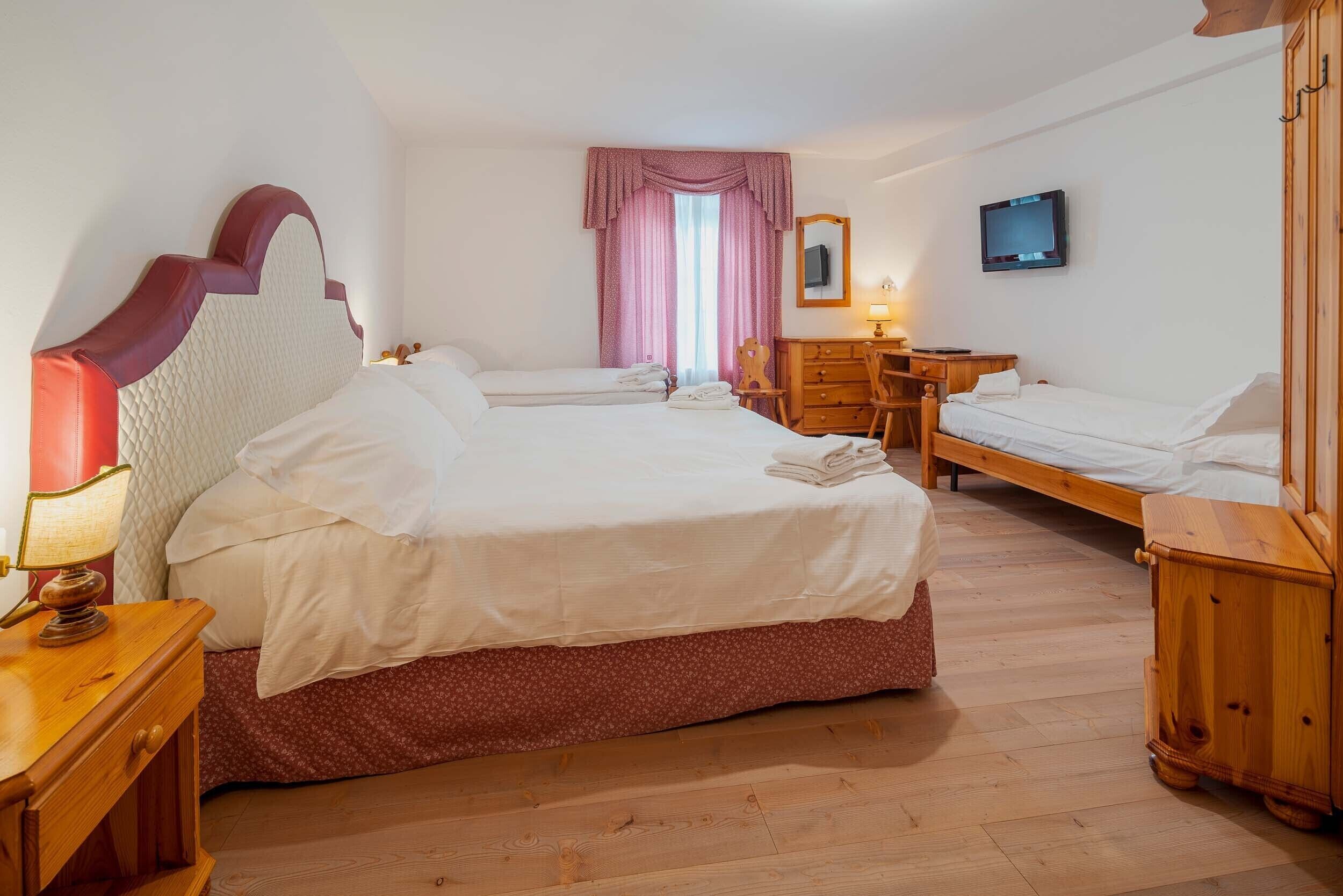 A hotel room with two single beds, a wooden dresser, a desk with a chair, a television, and a window with pink curtains.
