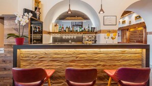 A bar area with a wooden counter, multiple liquor bottles, and three red chairs. - Hotel Royal (Cortina d'Ampezzo)