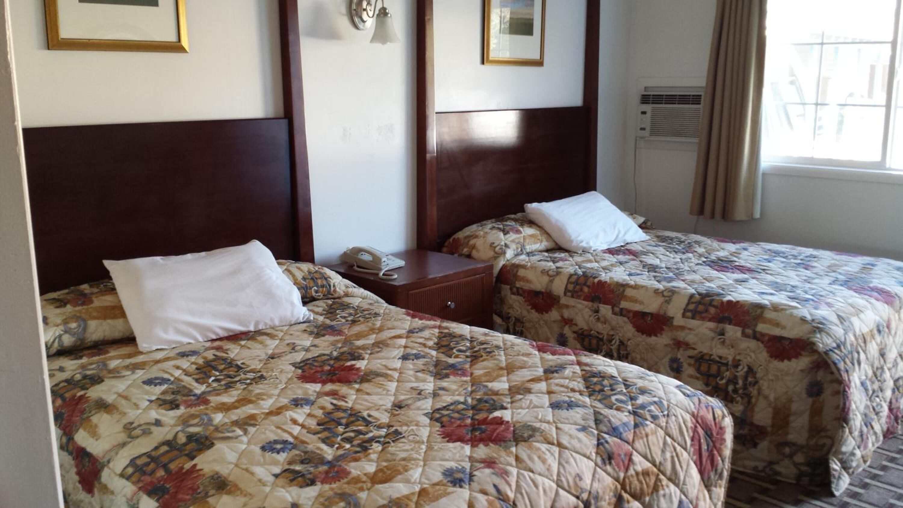 A hotel room with two beds, each with a floral quilt, a bedside table with a phone, and a wall-mounted light fixture.
