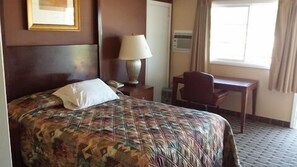 A hotel room with a bed, a desk, a chair, a lamp, and a window.