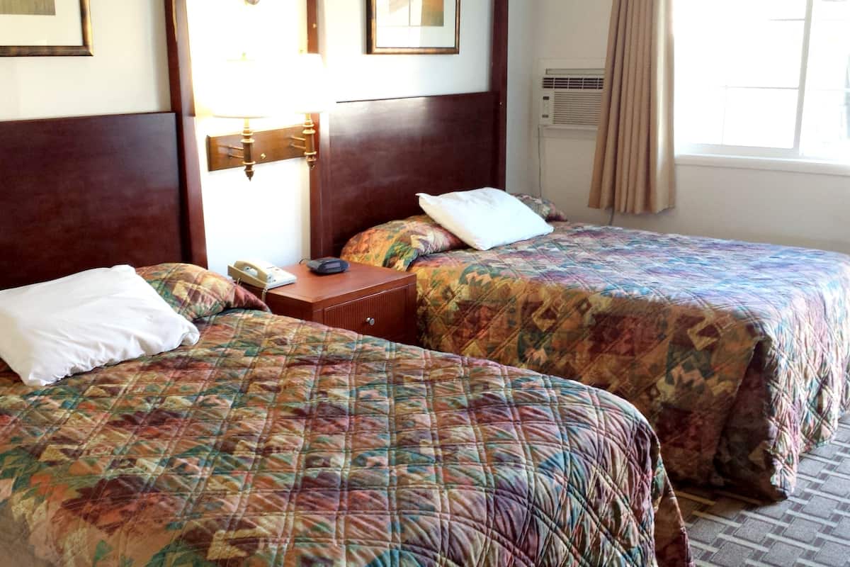 Habitación de hotel con dos camas, cada una con una colcha estampada y una almohada blanca. Entre las camas, un mesita de noche con una lámpara y un teléfono. La habitación tiene un cabecero de madera, una ventana con cortinas y un equipo de aire acondicionado en la pared.
