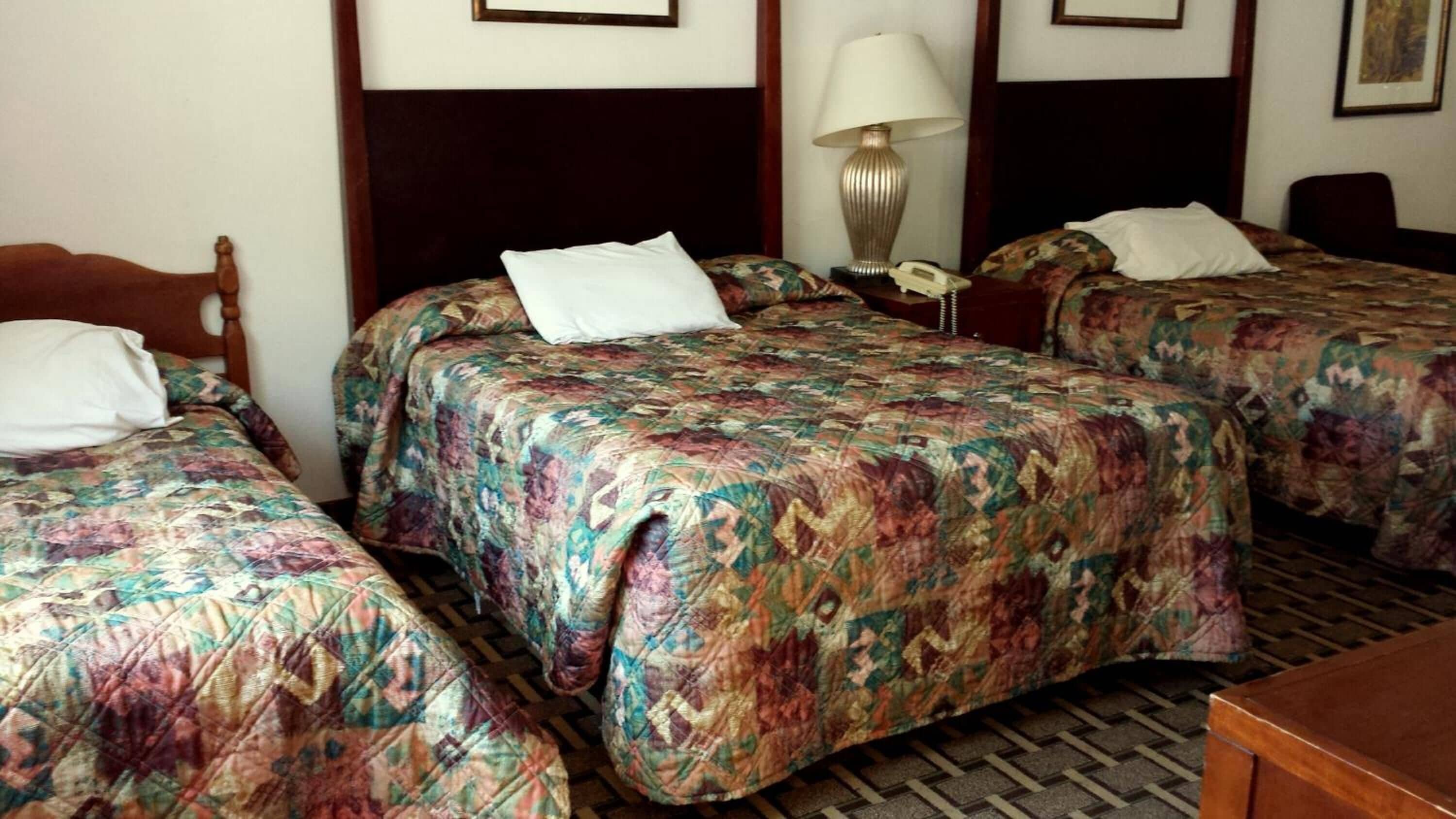 Een hotelkamer met drie enkele bedden, elk met een bloemenprint beddeken en een houten hoofdbord. Er staat een nachtkastje met een lamp en een telefoon. De kamer heeft een betegelde vloer en een houten bureau in de hoek.