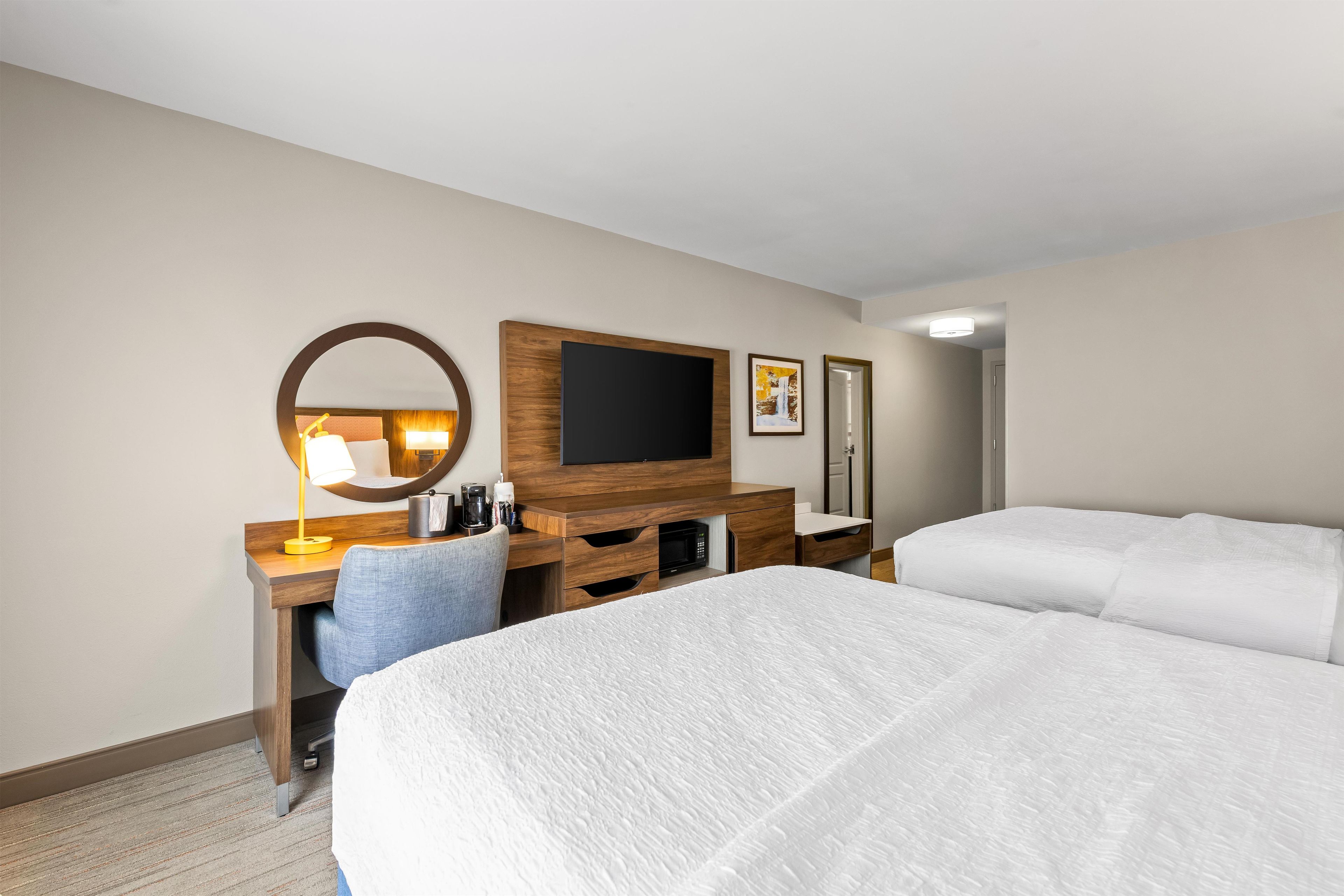 Quarto de hotel com cama, escrivaninha, cadeira, televisão, espelho e porta.