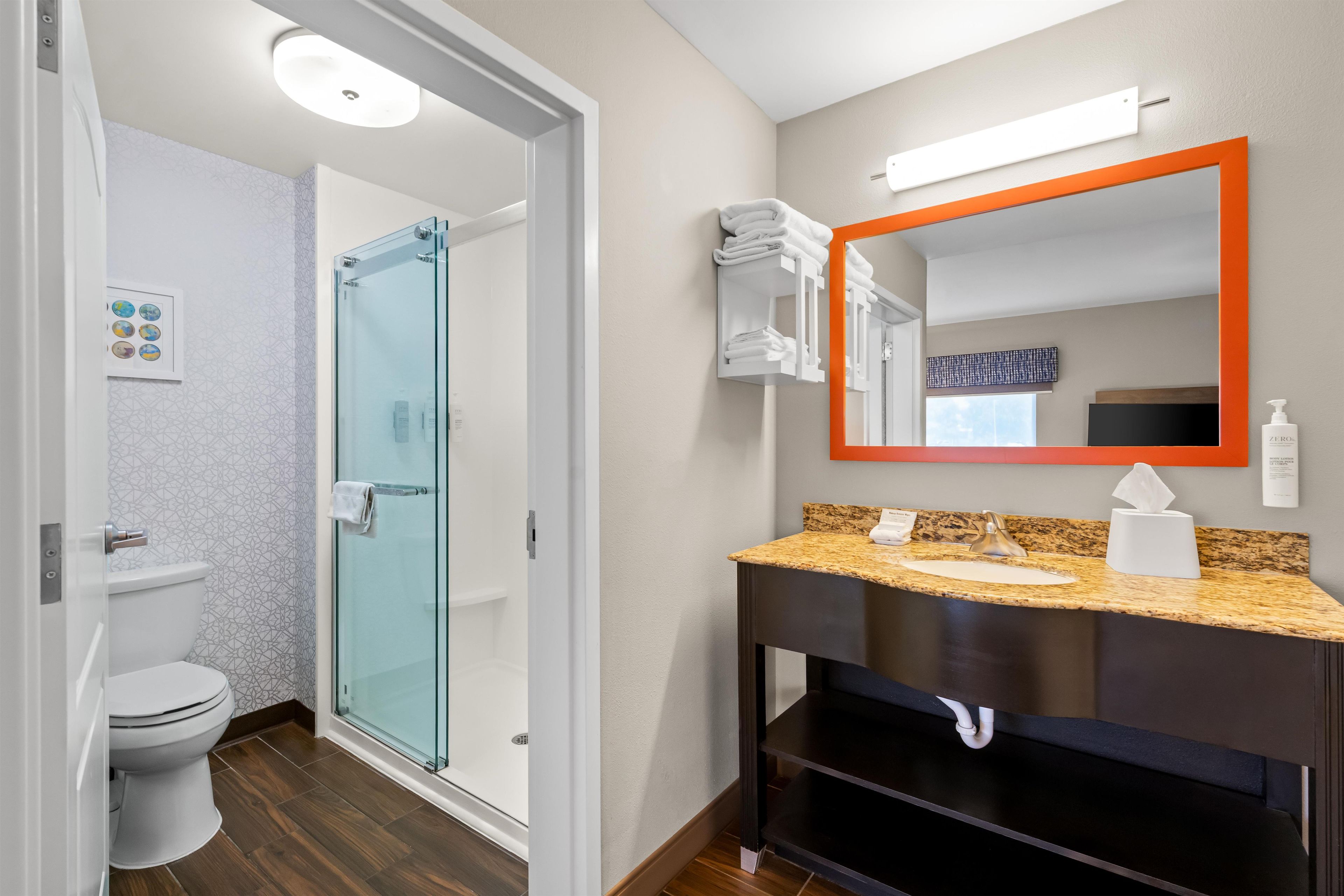 Un baño moderno con mampara de vidrio, inodoro blanco, tocador con encimera de granito y espejo con marco anaranjado.