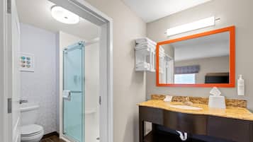 Un baño moderno con mampara de vidrio, inodoro blanco, tocador con encimera de granito y espejo con marco anaranjado.