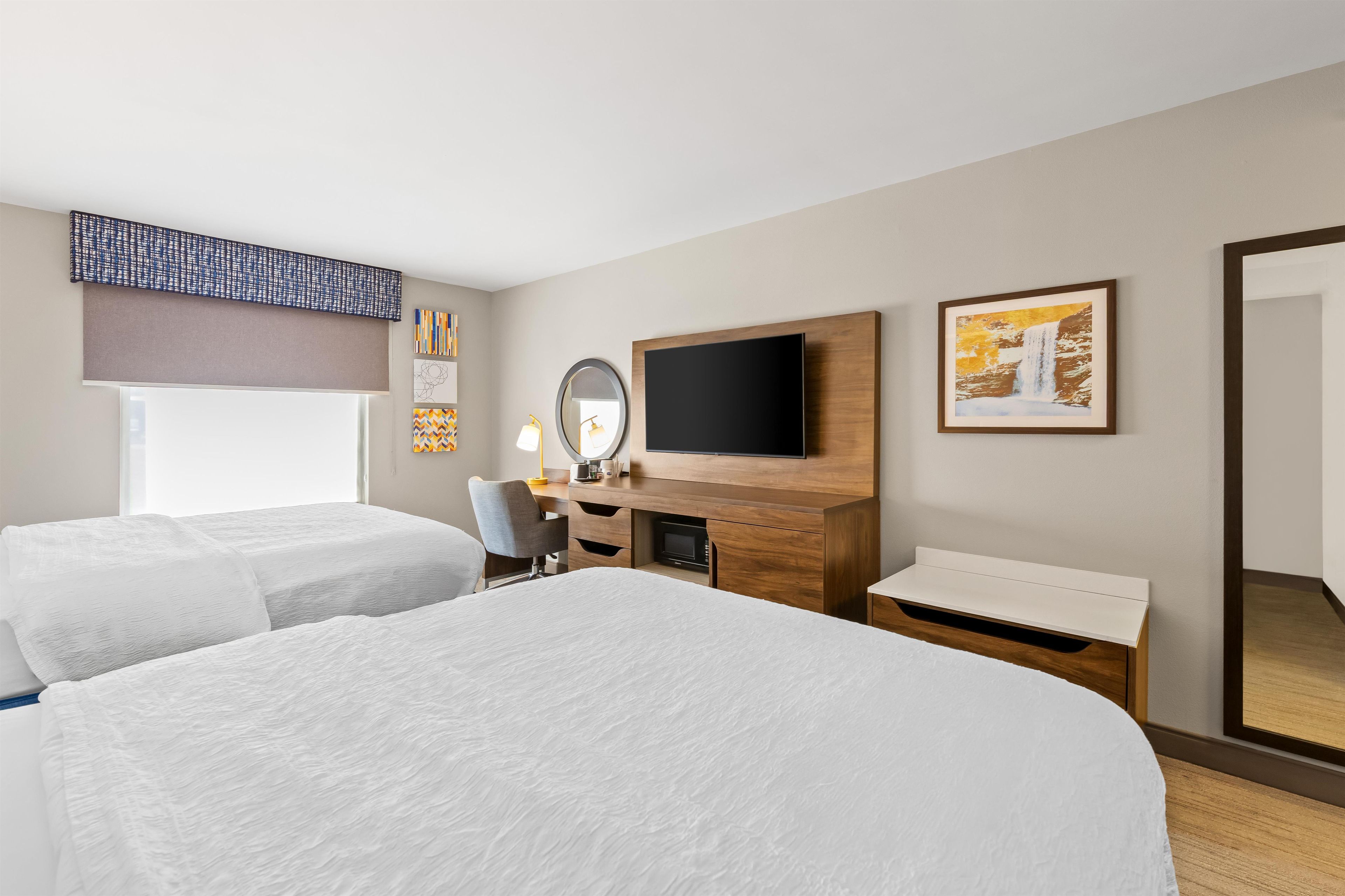 A hotel room with two beds, a desk with a flat-screen TV, a mirror, and artwork on the walls.