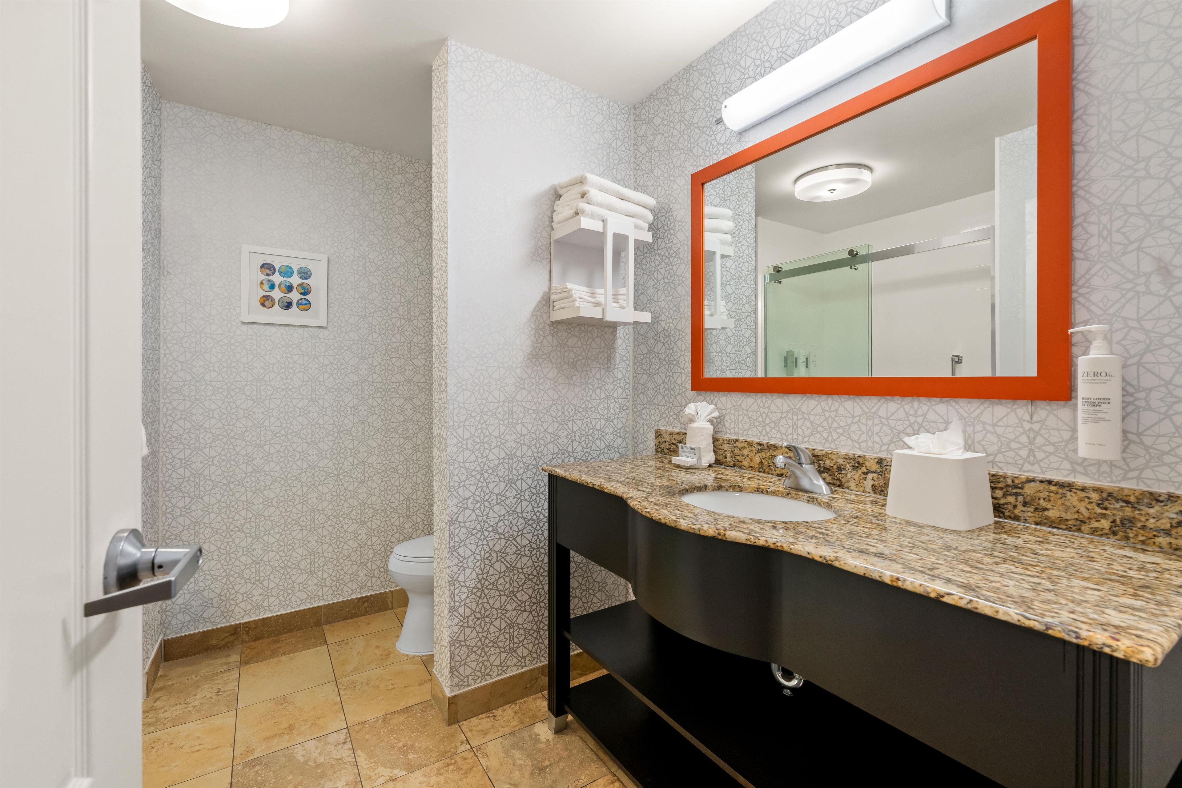 A modern bathroom with a granite countertop, a black sink, a mirror with an orange frame, and a shower area.