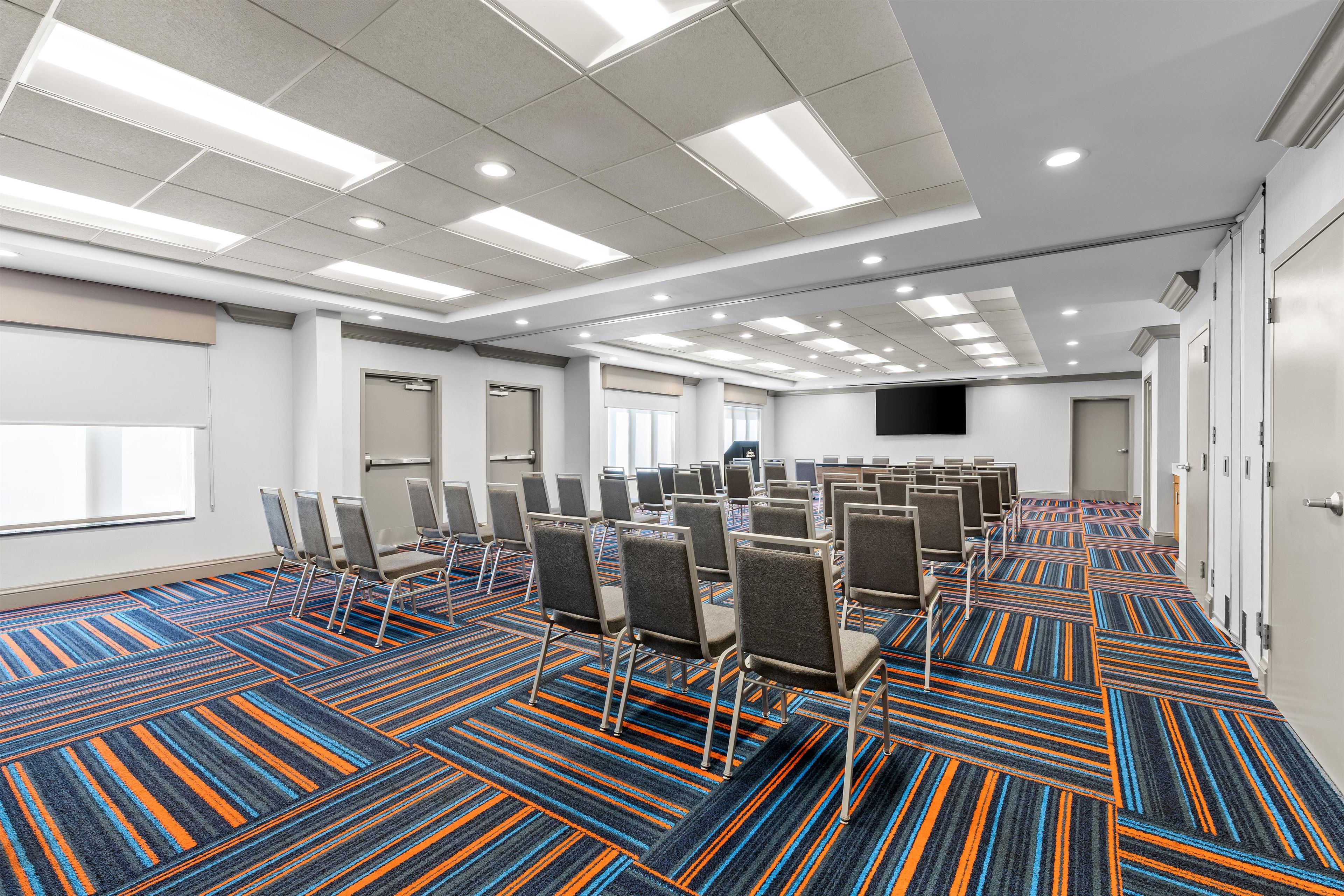 Un salón de conferencias con filas de sillas, piso alfombrado y una televisión de pantalla plana en la pared.