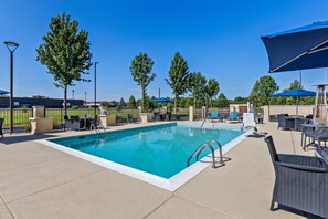 Una piscina rectangular con escalera metálica, rodeada de sillas de descanso y sombrillas, con árboles y un cielo despejado al fondo.