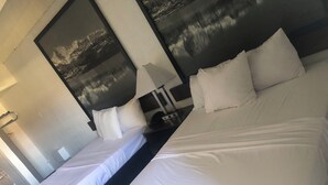 Habitación de hotel con dos camas, cada una con ropa y almohadas blancas. Hay una mesita de noche con una lámpara y un teléfono. La habitación tiene un ventanal con vistas a un paisaje montañoso.