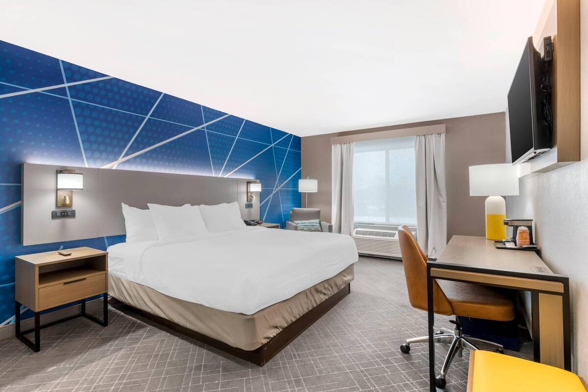 Une chambre d’hôtel avec un grand lit, un bureau avec une chaise, une table de chevet, un téléviseur fixé au mur et une fenêtre avec des rideaux.