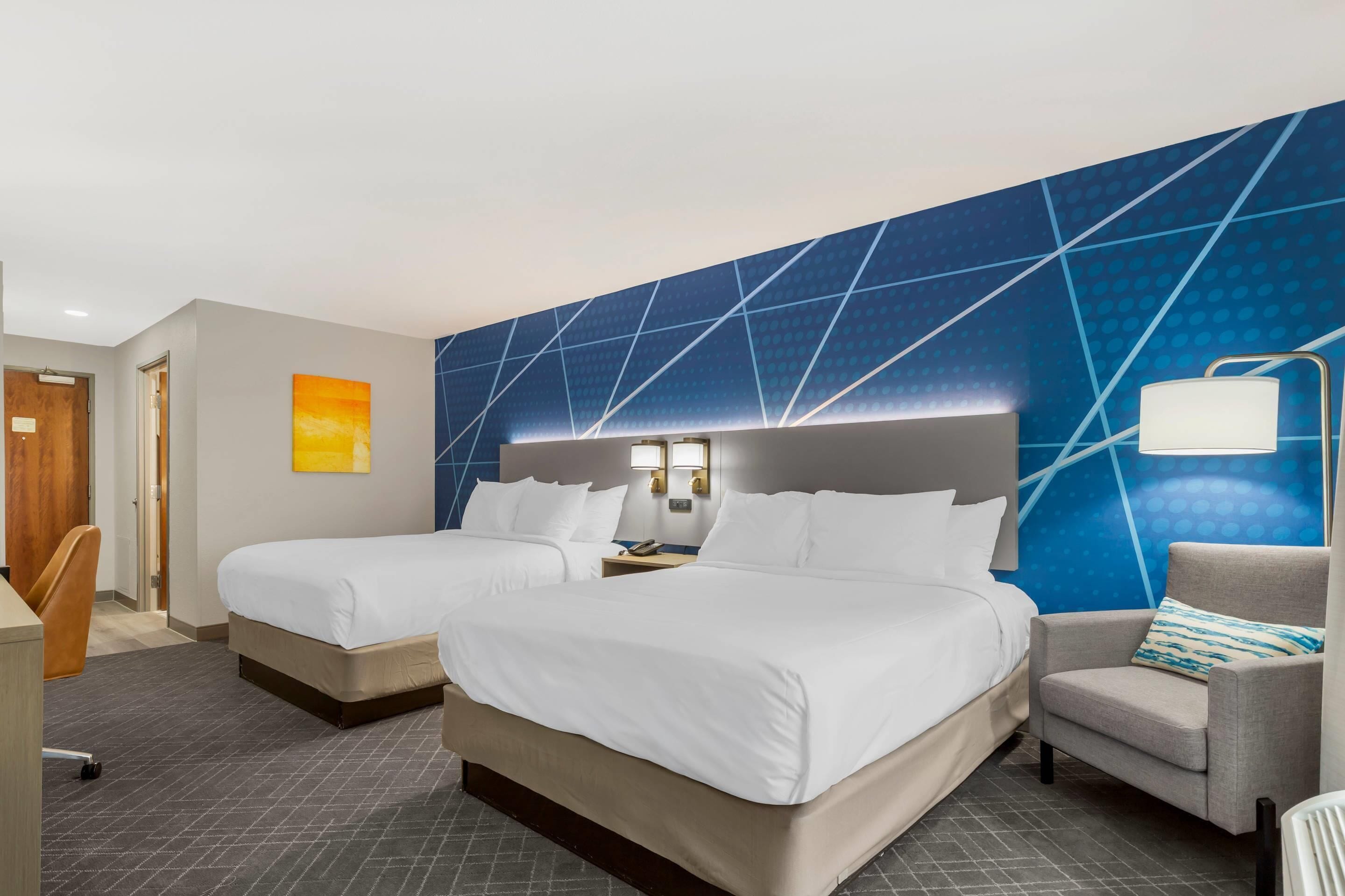 A hotel room with two beds, a desk, a chair, and a wall with a geometric pattern.