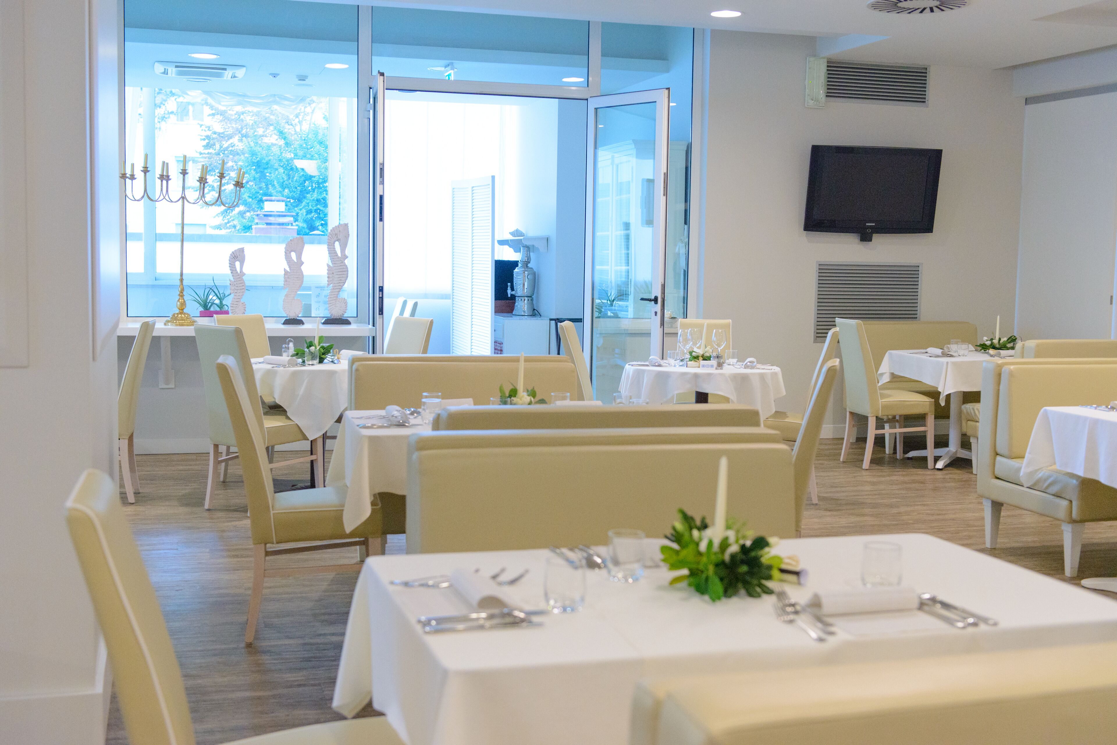 A dining area with multiple tables and chairs, a television mounted on the wall, and a view of the outdoors through large windows.