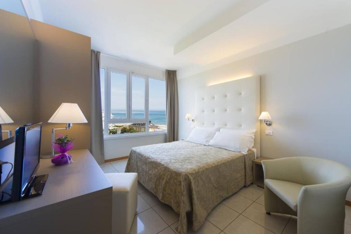 A hotel room with a bed, a desk with a computer, a chair, and a view of the sea.