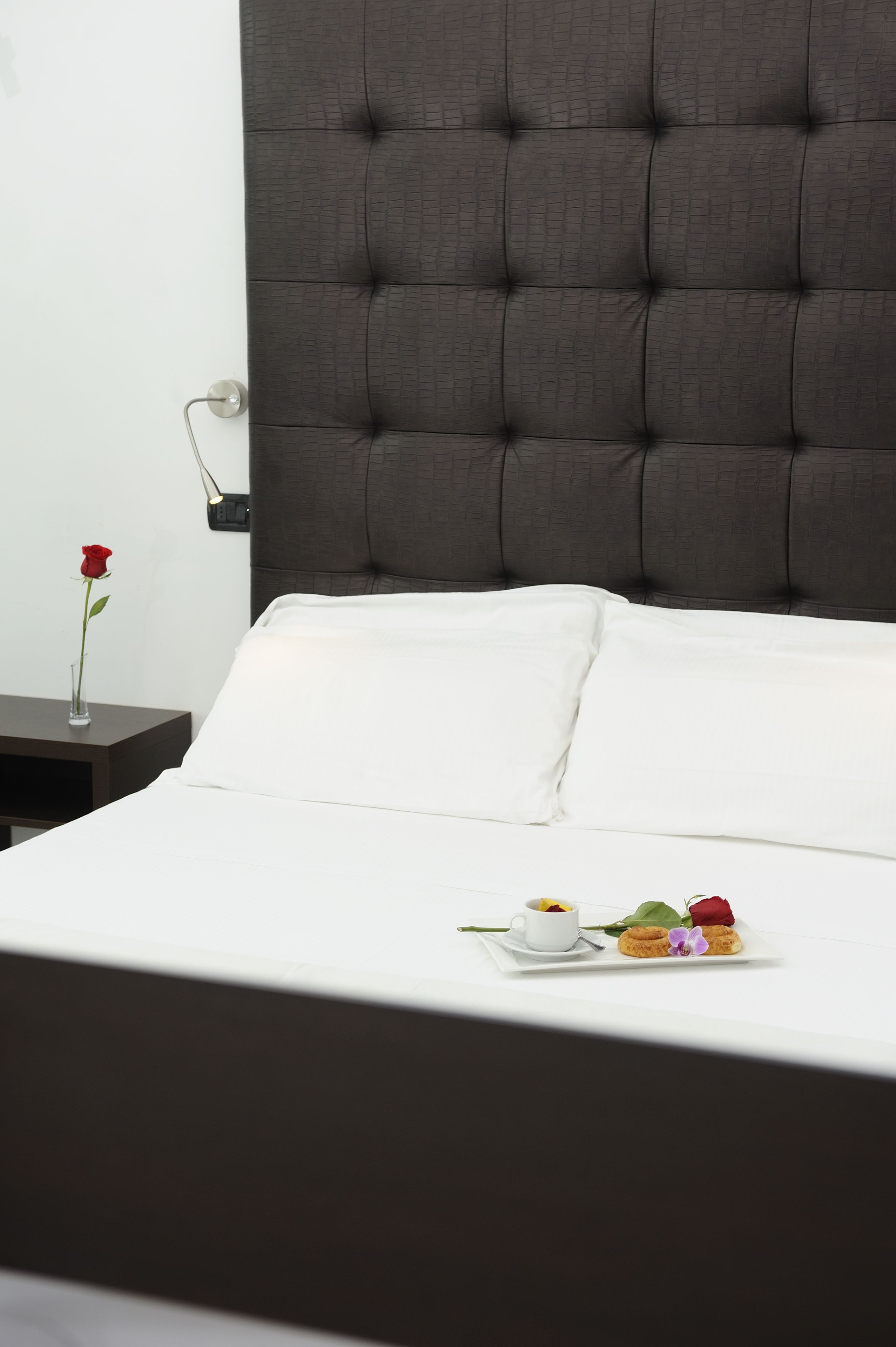 Una cama bien hecha con sábanas blancas y un cabecero oscuro. Una mesita de noche sostiene una rosa roja y una taza de café.