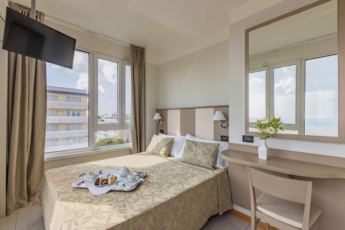 A hotel room with a bed, bedside lamps, a desk, a chair, a TV, and a window with a view of buildings.