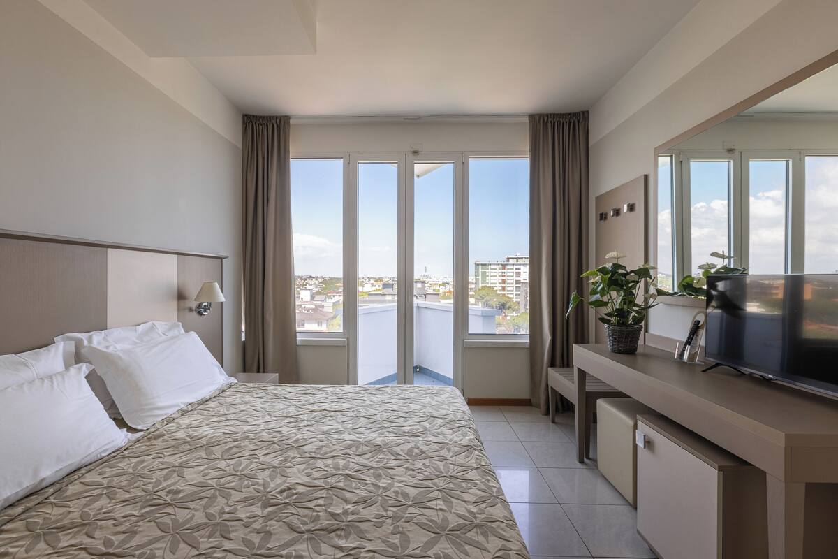 A hotel room with a bed, a desk with a TV, a potted plant, and a large window with curtains.