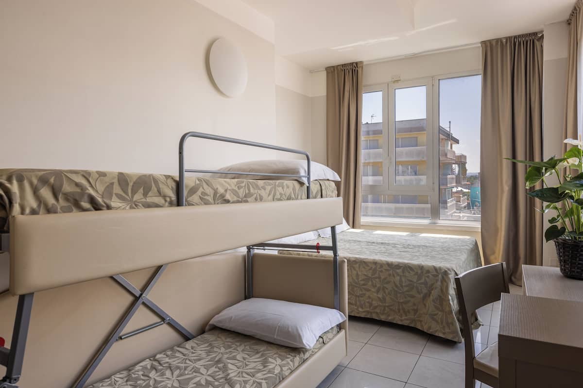 A hotel room with bunk beds, a dining table, a potted plant, and a window with a view of buildings.