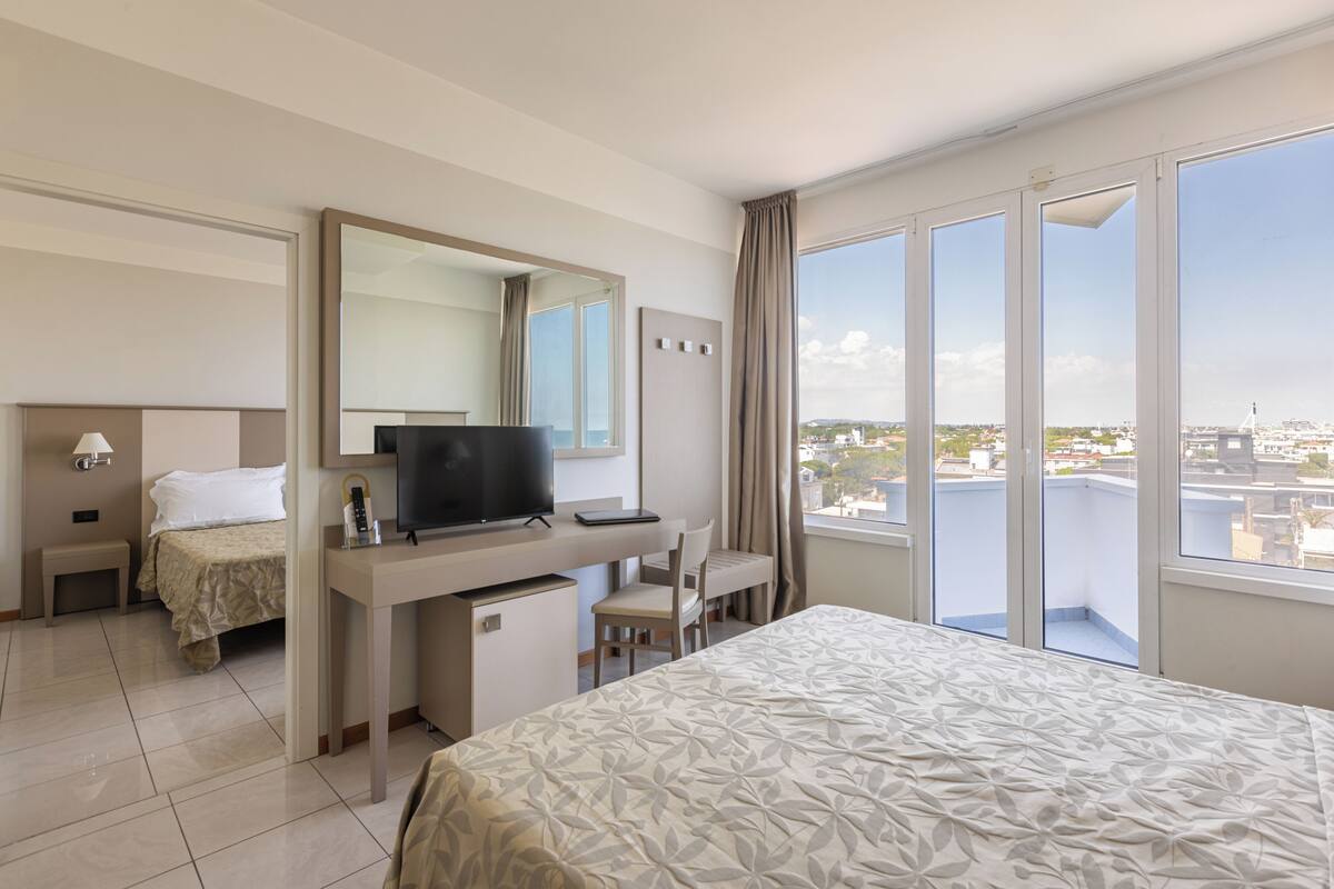 A hotel room with a bed, desk, chair, TV, and large windows offering a city view.