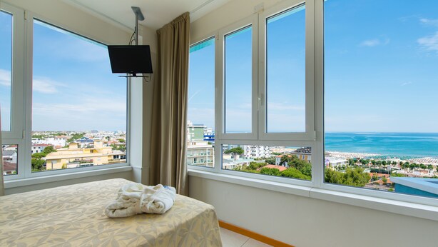 A hotel room with a bed, a TV, and large windows offering a view of the beach and cityscape. - Hotel Cristallo Riccione (Riccione)
