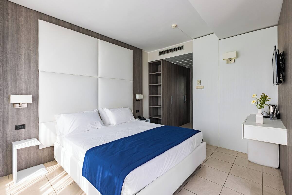 A modern bedroom with a large bed, a bedside table, a built-in wardrobe, a TV mounted on the wall, and a small desk with a vase.