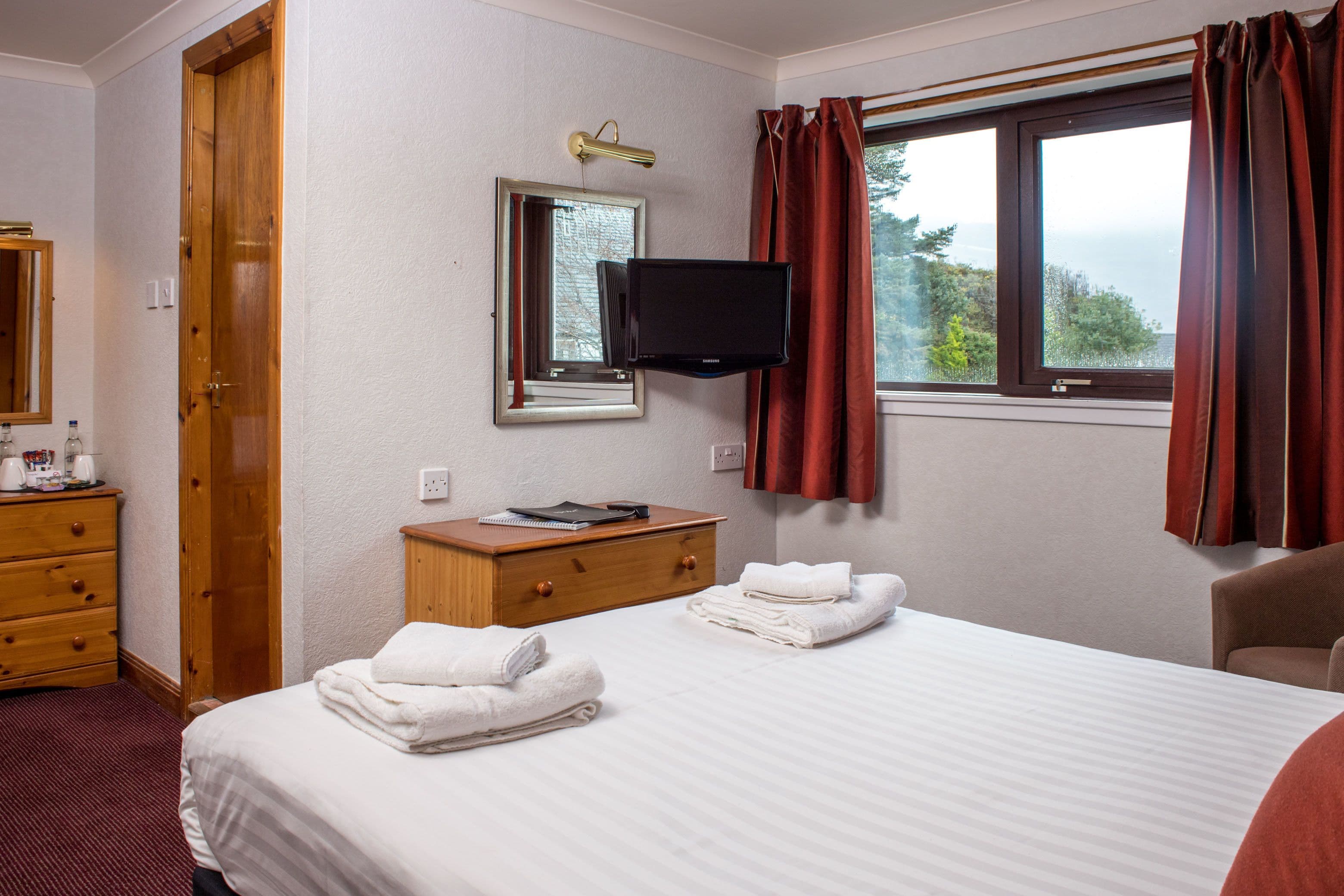 A hotel room with a bed, a TV, a wooden dresser, and a window with curtains.