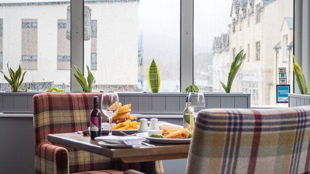 Ein Essbereich mit einem Tisch, gedeckt für zwei Personen. Es steht eine Weinflasche, ein Glas, ein Teller mit Essen und eine Wasserflasche darauf. Zwei Stühle mit Karomusterbezug sind vorhanden. Durch das Fenster hinter dem Tisch ist eine Aussicht auf Gebäude und eine Pflanze an der Fensterbank zu sehen.