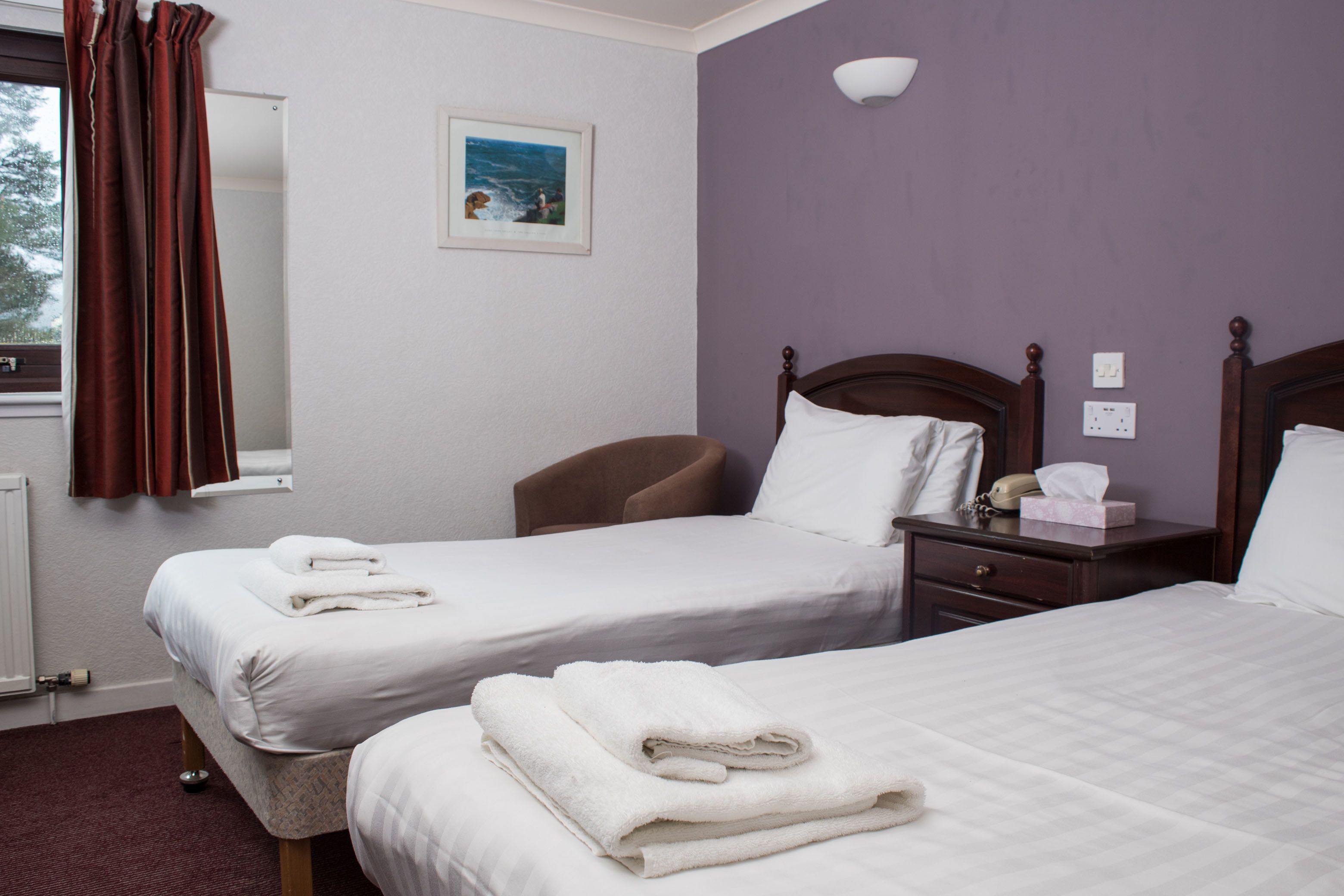 A hotel room with two beds, a window with curtains, a bedside table with a lamp, and a painting on the wall.