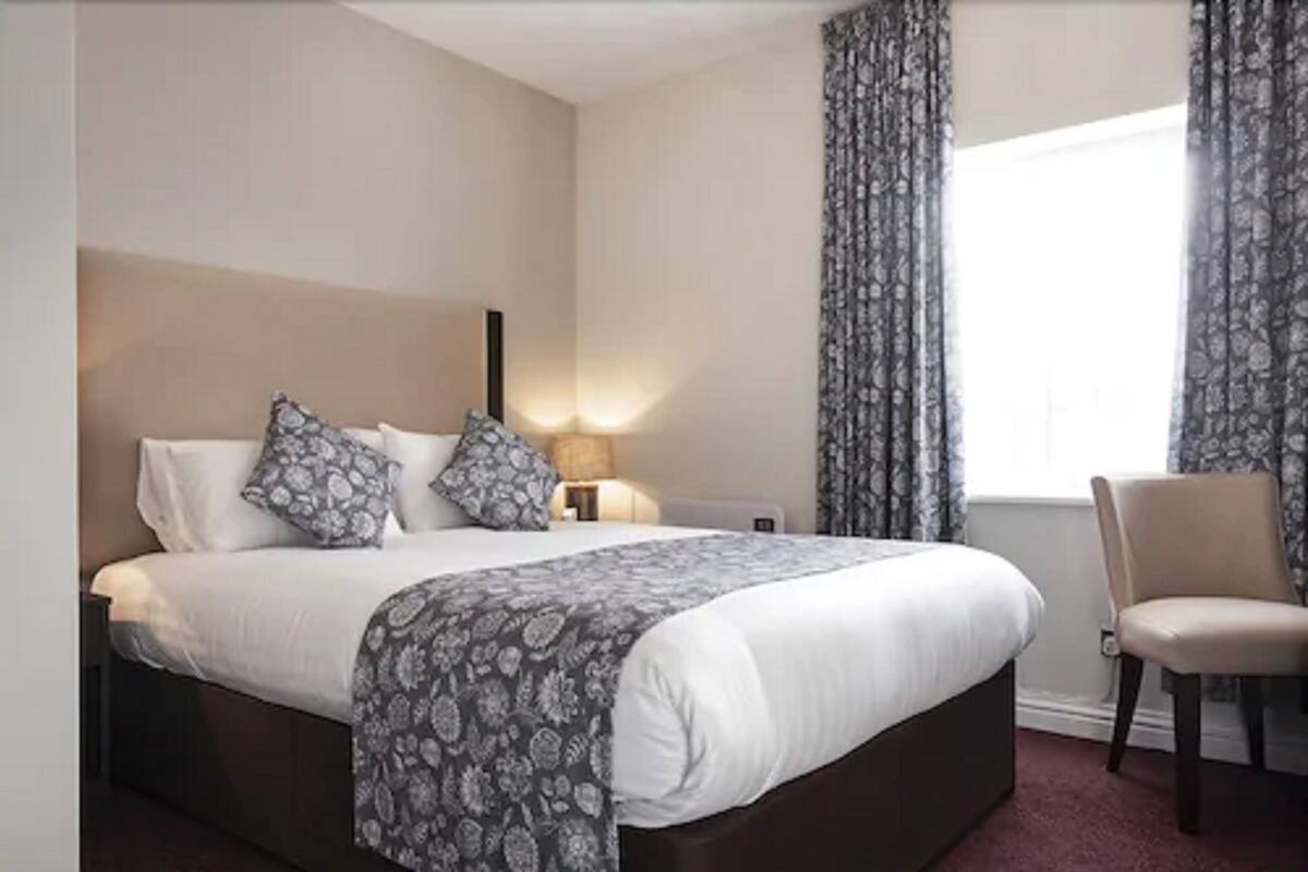 A hotel room with a large bed, a bedside table with a lamp, a chair, and a window with curtains.