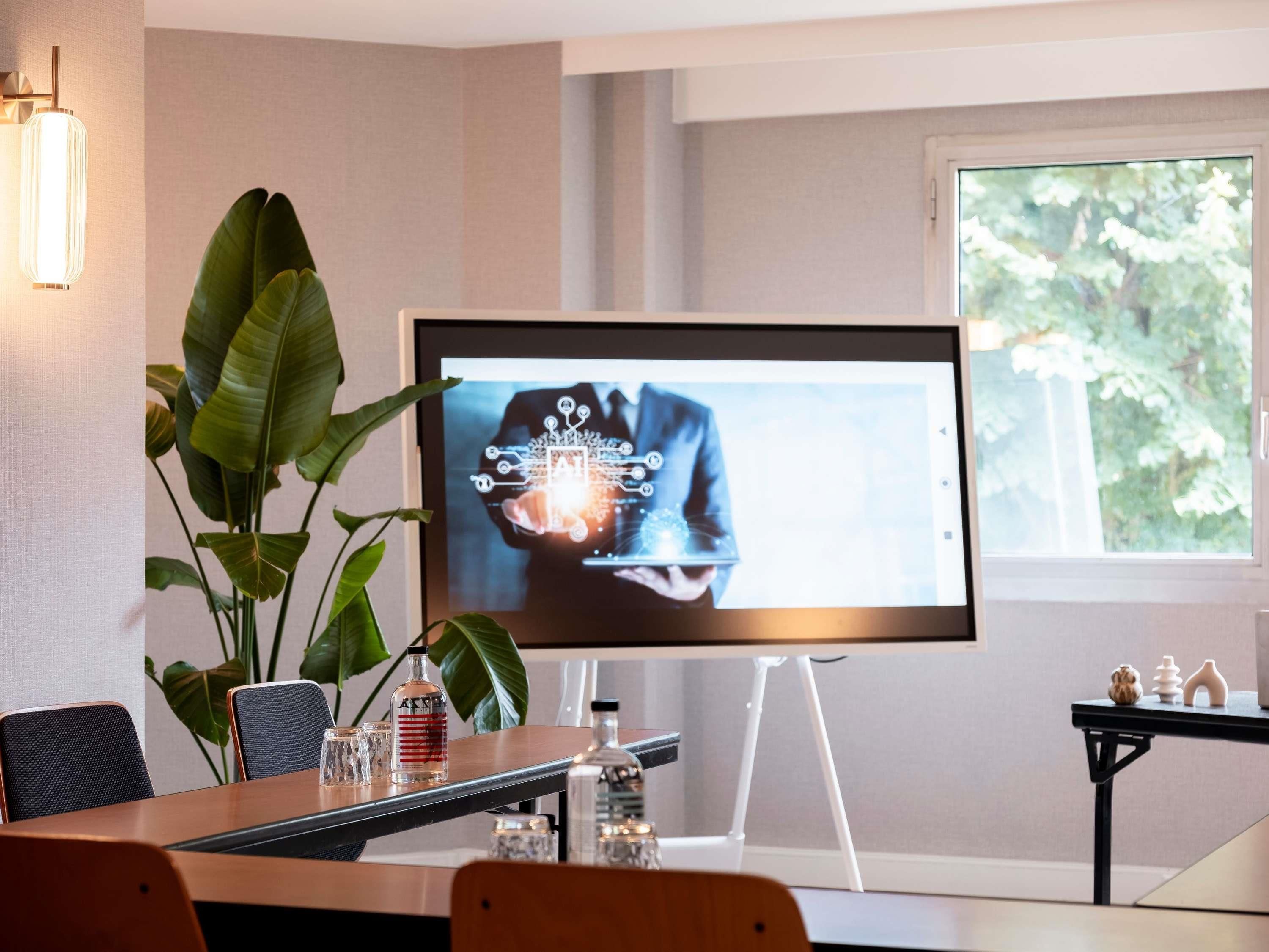 Een vergaderruimte met een groot scherm waarop een digitale grafiek wordt getoond, een houten tafel met stoelen, een potplant en een raam met uitzicht op bomen.