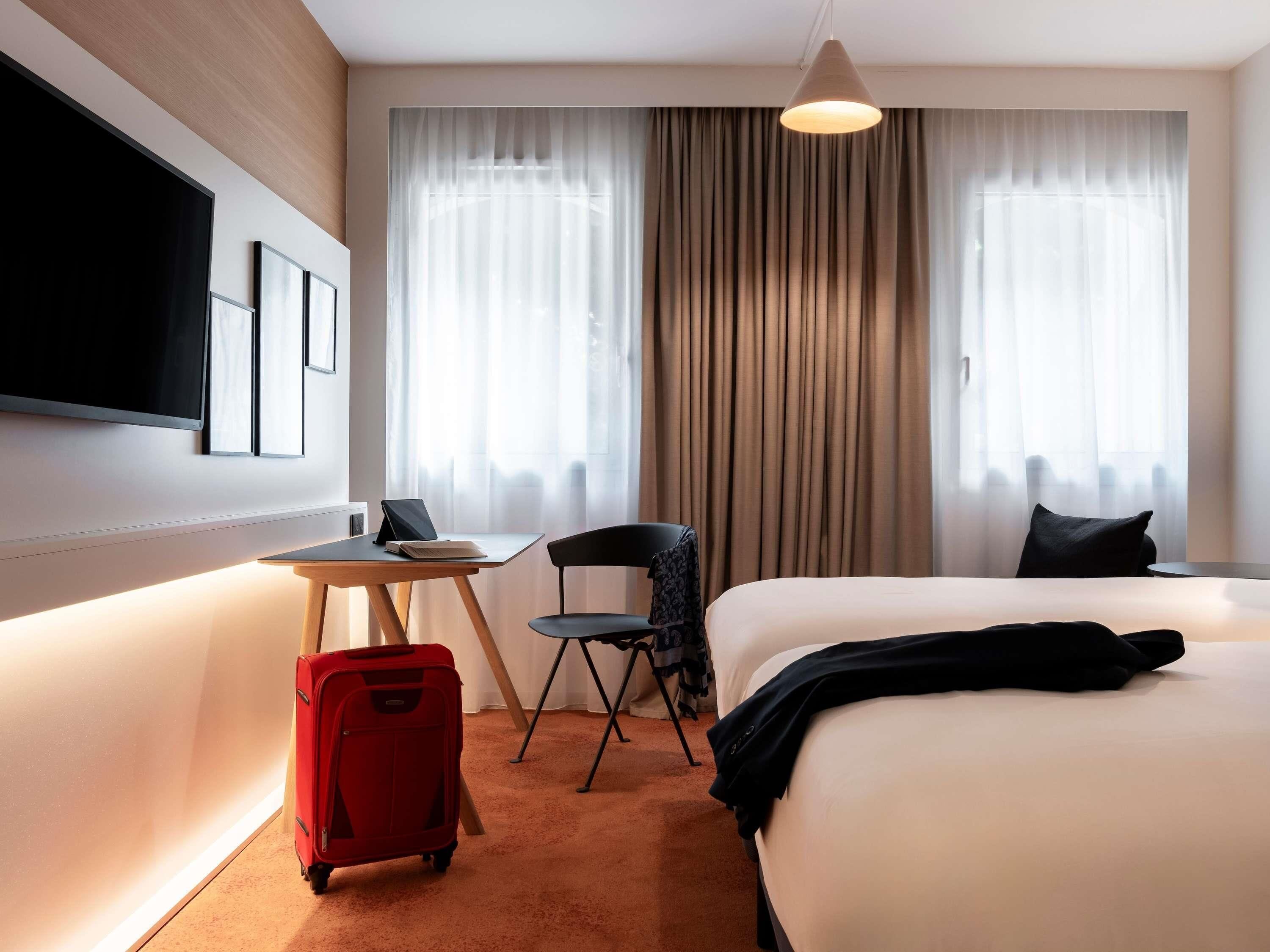 Une chambre d’hôtel avec un lit, un bureau et une chaise, une valise rouge et un téléviseur fixé au mur.