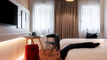 Une chambre d’hôtel avec un lit, un bureau et une chaise, une valise rouge et un téléviseur fixé au mur.