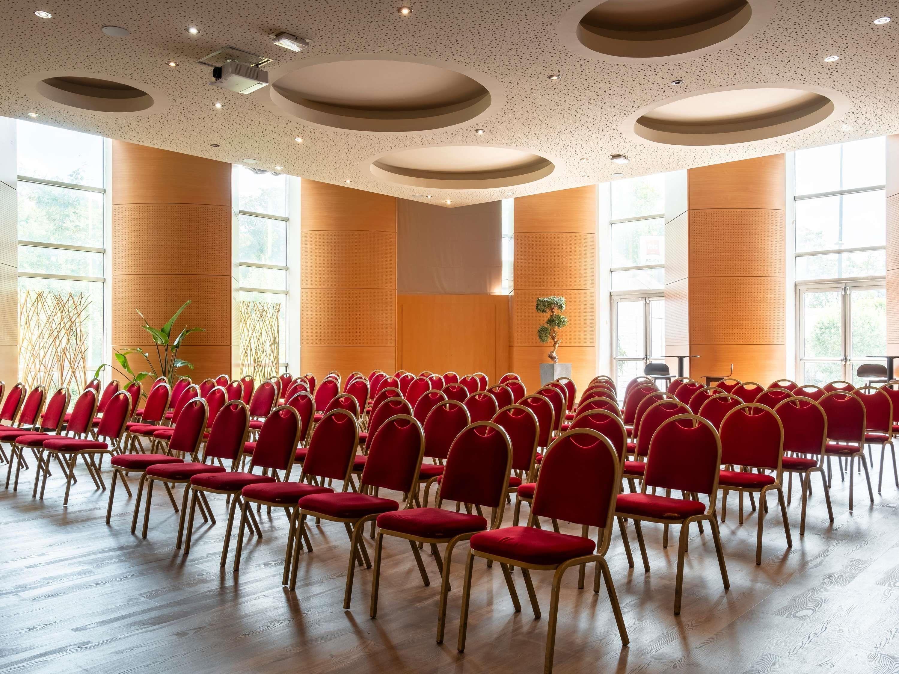 Un salón de conferencias con filas de sillas rojas, suelo de madera y amplias ventanas.