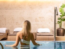 Una persona con traje de baño blanco sentada junto a una piscina, con sillas de descanso y una planta en maceta al fondo.