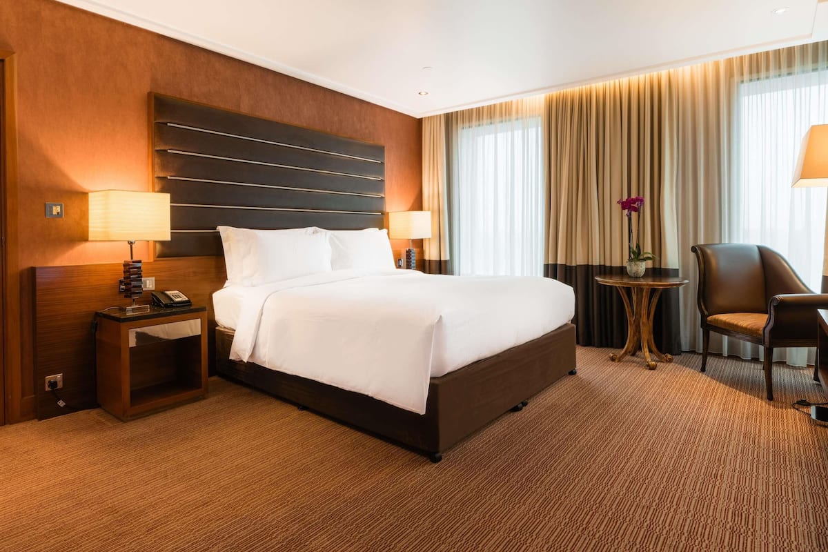 A hotel room with a large bed, bedside lamps, a small table with a plant, a chair, and a view of the outside through curtains.