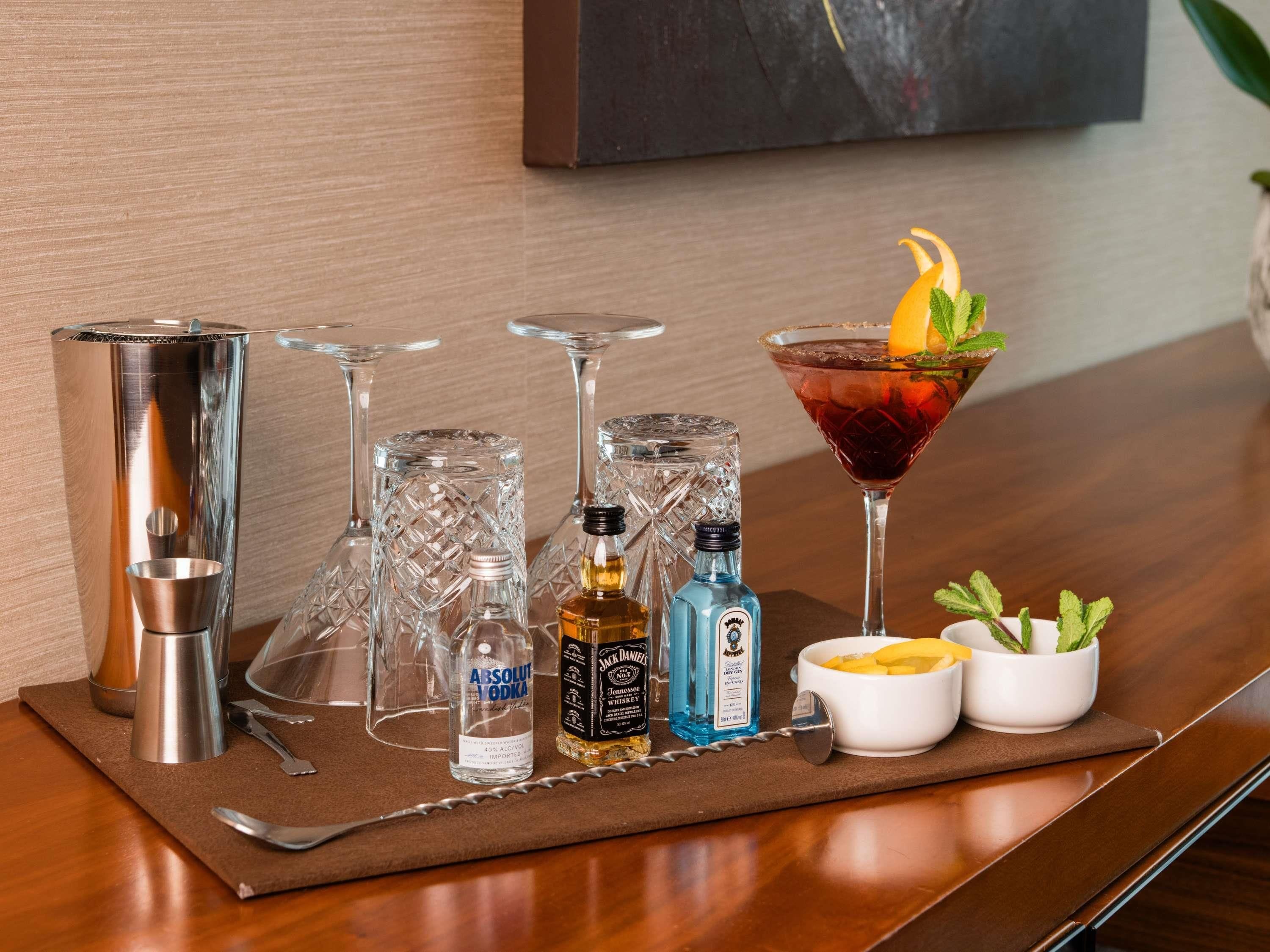 A bar setup with a martini glass, a cocktail shaker, a bottle of Absolut Vodka, a bottle of Jack Daniel's Whiskey, and a bowl of fruit on a wooden table.