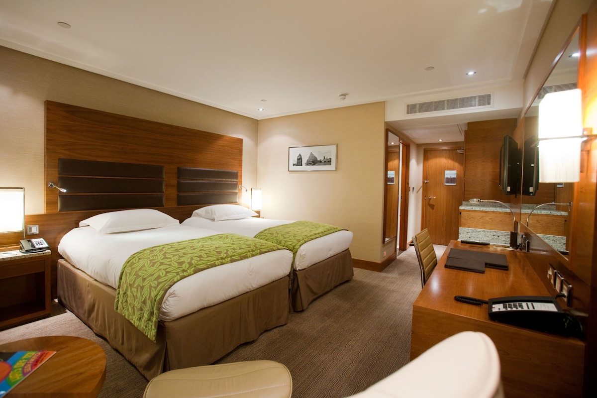 A hotel room with two beds, a desk with a keyboard, a chair, a bedside table with a lamp, and a picture on the wall.