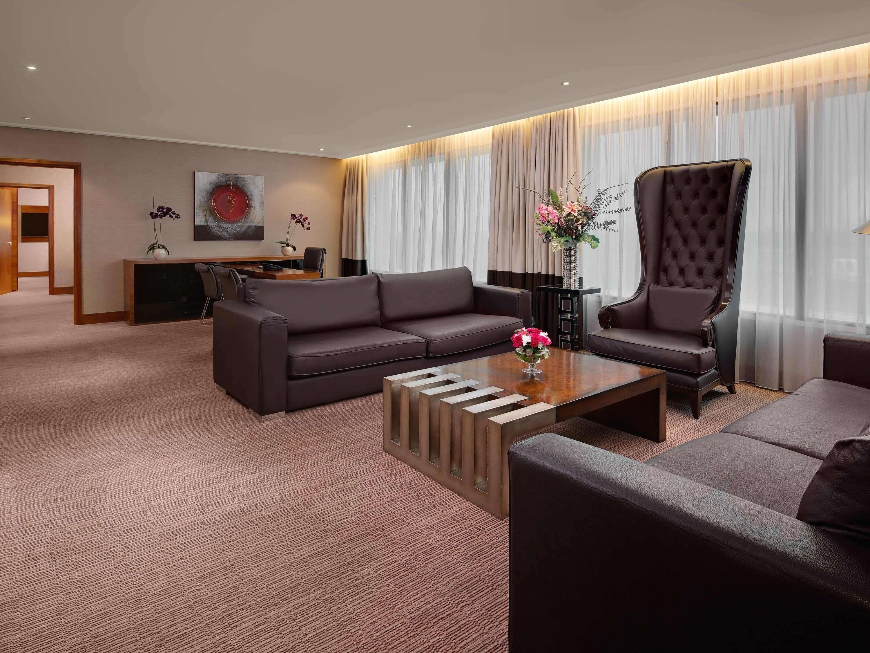 A modern hotel room with a dark brown leather sofa, a matching armchair, a wooden coffee table, and a desk with a chair.