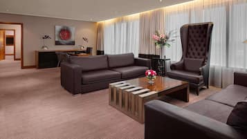 A modern hotel room with a dark brown leather sofa, a matching armchair, a wooden coffee table, and a desk with a chair.