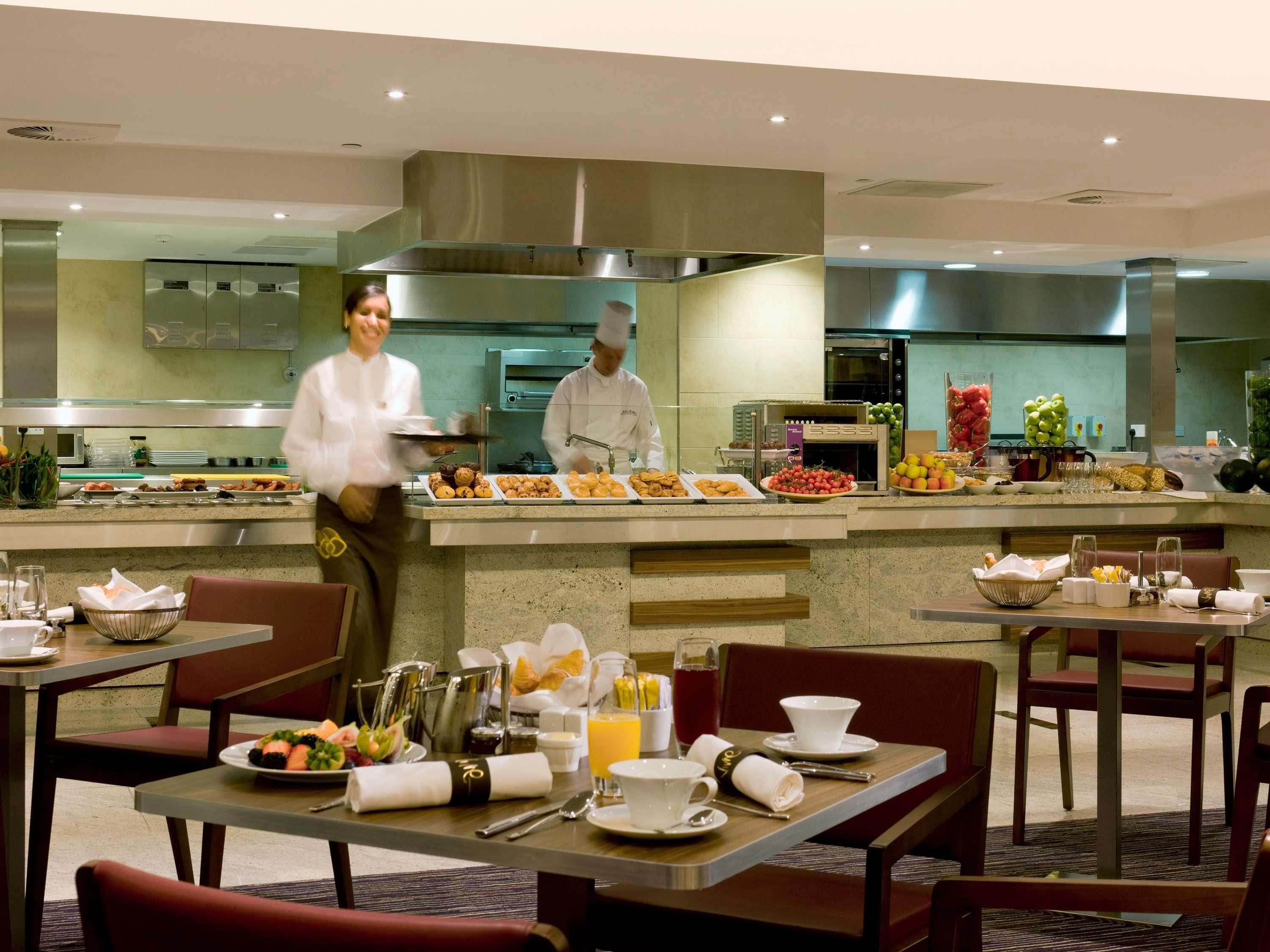 A hotel dining area with tables set for breakfast, a buffet with various food items, and a kitchen area in the background.