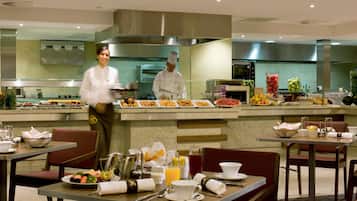 A hotel dining area with tables set for breakfast, a buffet with various food items, and a kitchen area in the background.