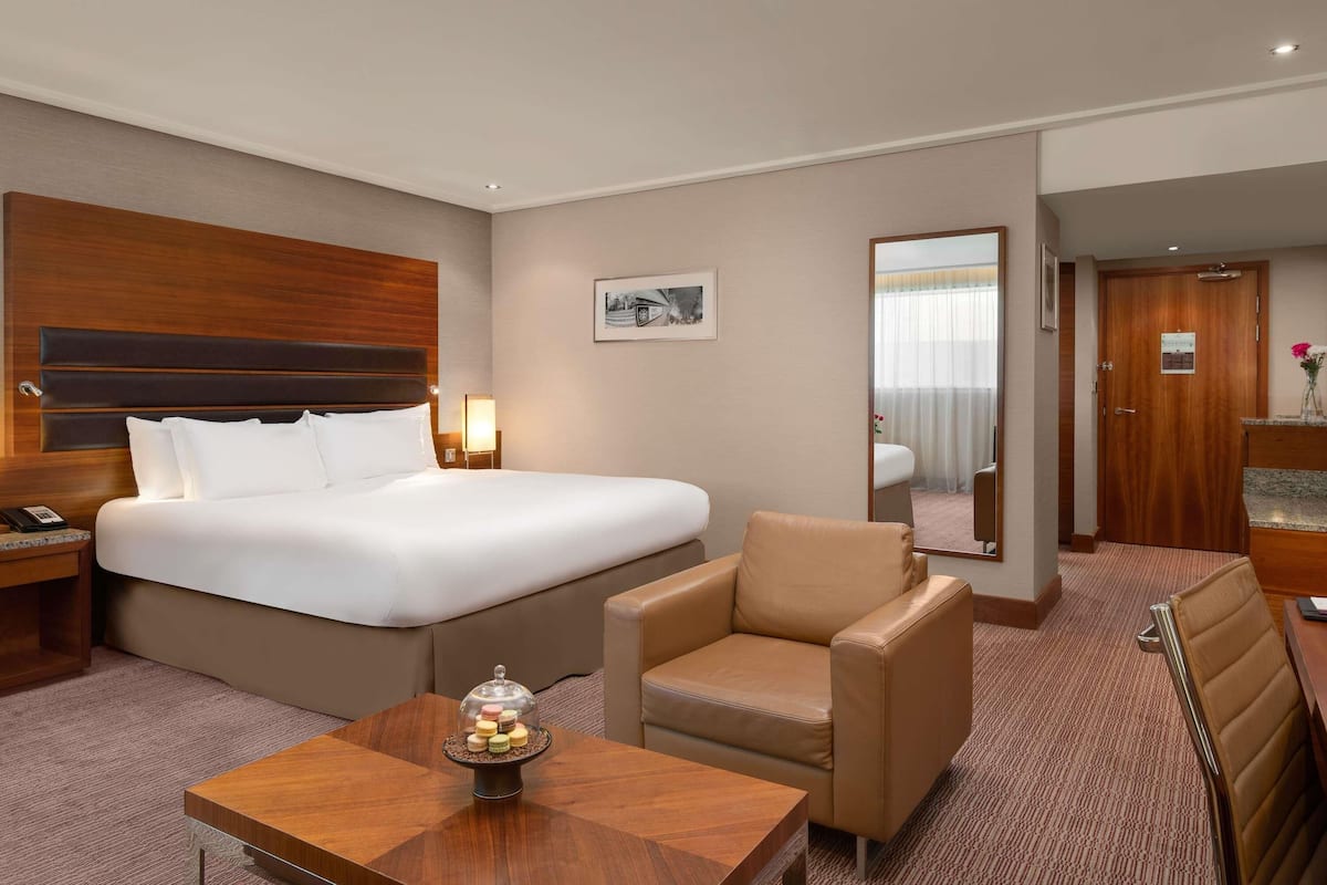 A hotel room with a large bed, a small armchair, a wooden table with a decorative item, a nightstand, and a mirror.