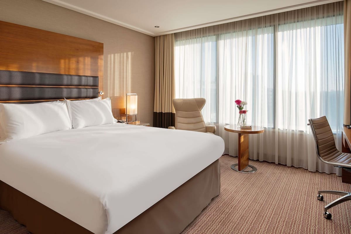 A hotel room with a large bed, a desk with a chair, a small table with flowers, and a view of the city through sheer curtains.