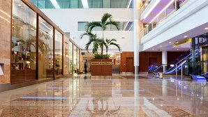 Een ruime luchthaventerminal met een gepolijste marmeren vloer, glazen etalages en een centrale plantenbak met palmboompjes.