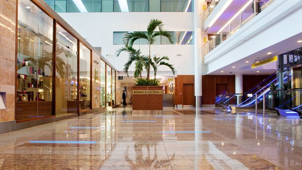 A spacious airport terminal with a polished marble floor, glass storefronts, and a central planter with palm trees. - Sofitel London Heathrow (West Drayton)